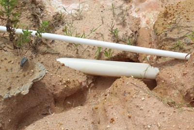 A broken white plastic pipe lies partially buried in sandy soil, surrounded by small rocks and sparse vegetation. The sandy ground features various shades and textures, with some exposed rocky surfaces.