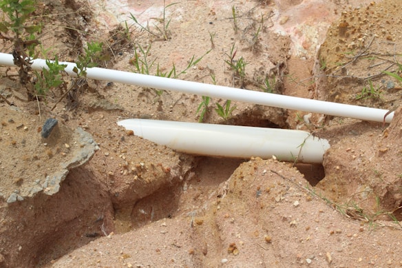 A broken white plastic pipe lies partially buried in sandy soil, surrounded by small rocks and sparse vegetation. The sandy ground features various shades and textures, with some exposed rocky surfaces.