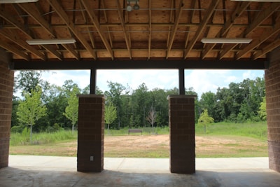 A covered pavilion structure with a wooden ceiling and exposed beams is seen from the inside. There are three brick pillars in the foreground, and beyond them is a grassy field with a few small trees against a backdrop of dense woods. A solitary bench is situated in the open space, providing a focal point amidst the lush greenery.