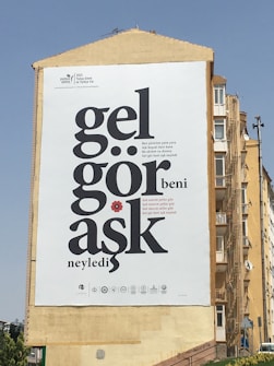 A large poster is mounted on the side of a building featuring bold black text with a red flower detail. The text includes quotes and several logos along the bottom. The building is against a clear blue sky.