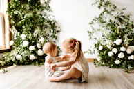 Two young children are sitting on a wooden floor in front of a wall adorned with green foliage and white flowers. The older child, a girl with a ponytail and wearing a dress with a floral pattern, appears to be talking to or playing with the younger child, a baby. Natural light is streaming in from a window on the left side.
