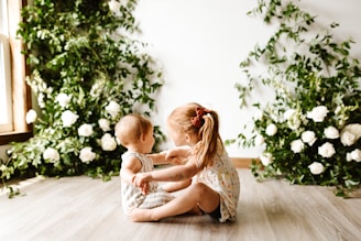 Two young children are sitting on a wooden floor in front of a wall adorned with green foliage and white flowers. The older child, a girl with a ponytail and wearing a dress with a floral pattern, appears to be talking to or playing with the younger child, a baby. Natural light is streaming in from a window on the left side.