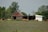 A rural scene with a weathered wooden barn and a white shed surrounded by lush green trees. Several brown cows graze in an open field speckled with small wildflowers.