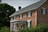 A two-story brick house with white trim features several windows with dark shutters, a pitched roof, and an ornate white porch with decorative elements. Lush greenery surrounds the house, including bushes and leafy trees, suggesting a serene and traditional setting.