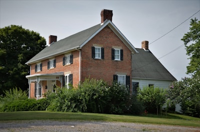 A traditional two-story brick house with black shutters and a gable roof. Surrounded by lush greenery and trees, the property includes a manicured lawn and shrubbery, with a gravel path leading up to the house. The architecture is vintage and robust, evoking a sense of history and permanence.