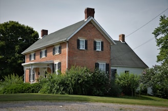 A traditional two-story brick house with black shutters and a gable roof. Surrounded by lush greenery and trees, the property includes a manicured lawn and shrubbery, with a gravel path leading up to the house. The architecture is vintage and robust, evoking a sense of history and permanence.