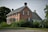 A traditional two-story brick house with black shutters and a gable roof. Surrounded by lush greenery and trees, the property includes a manicured lawn and shrubbery, with a gravel path leading up to the house. The architecture is vintage and robust, evoking a sense of history and permanence.