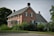 A traditional two-story brick house with black shutters and a gable roof. Surrounded by lush greenery and trees, the property includes a manicured lawn and shrubbery, with a gravel path leading up to the house. The architecture is vintage and robust, evoking a sense of history and permanence.