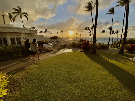 A scenic landscape featuring a lush green lawn leading to the ocean, with a sunset casting warm golden light across the scene. Palm trees are silhouetted against the sky, and two people walk along a path on the left. The building to the left has a rounded architecture, adding a modern touch to the tropical setting.