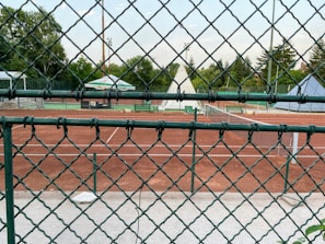 A sports scene captured through a chain-link fence, focusing on a tennis court with a red clay surface. The court is empty, surrounded by greenery and tall trees in the background. A large umbrella provides shade, and tennis nets are visible.