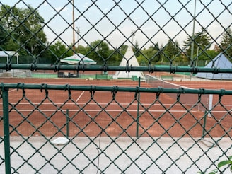 A sports scene captured through a chain-link fence, focusing on a tennis court with a red clay surface. The court is empty, surrounded by greenery and tall trees in the background. A large umbrella provides shade, and tennis nets are visible.