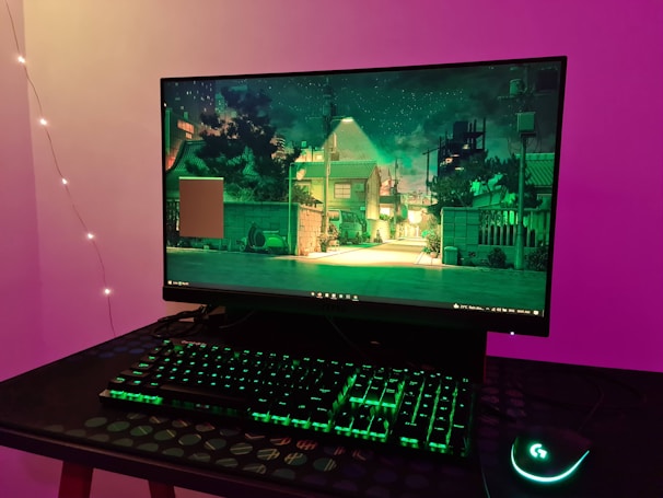 A computer setup is on display, featuring a monitor showcasing a scene of a serene, illuminated urban street at night with trees and houses. The setup includes a backlit keyboard and a gaming mouse with green lighting, all placed on a textured table. The background wall is illuminated with purple and ambient lighting from string lights.