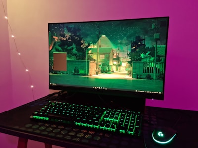A computer setup is on display, featuring a monitor showcasing a scene of a serene, illuminated urban street at night with trees and houses. The setup includes a backlit keyboard and a gaming mouse with green lighting, all placed on a textured table. The background wall is illuminated with purple and ambient lighting from string lights.