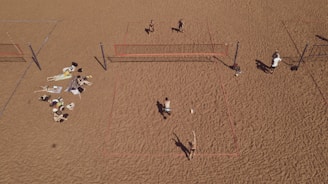 An aerial view of a sandy beach area featuring multiple beach volleyball courts with players actively engaged in a game. On the left side, a group is relaxing on towels, sunbathing and socializing. The sand is uniformly spread, creating a wide open space.