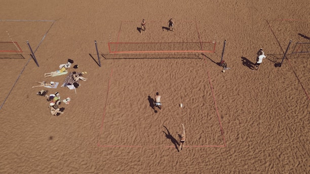 An aerial view of a sandy beach area featuring multiple beach volleyball courts with players actively engaged in a game. On the left side, a group is relaxing on towels, sunbathing and socializing. The sand is uniformly spread, creating a wide open space.