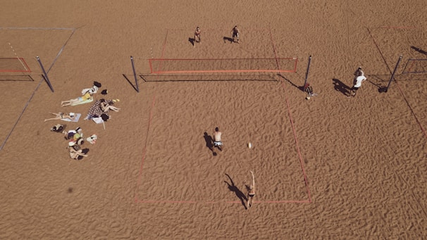 An aerial view of a sandy beach area featuring multiple beach volleyball courts with players actively engaged in a game. On the left side, a group is relaxing on towels, sunbathing and socializing. The sand is uniformly spread, creating a wide open space.
