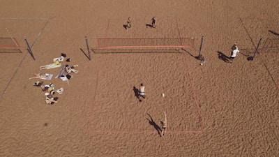 An aerial view of a sandy beach area featuring multiple beach volleyball courts with players actively engaged in a game. On the left side, a group is relaxing on towels, sunbathing and socializing. The sand is uniformly spread, creating a wide open space.