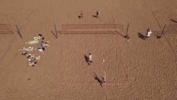 An aerial view of a sandy beach area featuring multiple beach volleyball courts with players actively engaged in a game. On the left side, a group is relaxing on towels, sunbathing and socializing. The sand is uniformly spread, creating a wide open space.