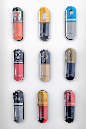 Nine large capsule-shaped objects are arranged in a grid pattern, each featuring different brand logos and vibrant colors. The brands include 7-Eleven, Snapchat, PayPal, Cartier, Dolce & Gabbana, Yves Saint Laurent, Marc Jacobs, In-N-Out Burger, and Tinder. Each capsule is labeled with '100MG' at the bottom.