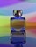 A glass perfume bottle labeled 'ECLAT mademoiselle' from Oriflame Sweden is prominently displayed, standing on a reflective surface. The background features a gradient of vibrant colors including purple, blue, green, yellow, and orange, creating a colorful and artistic setting.