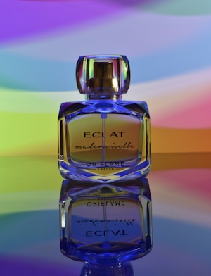 A glass perfume bottle labeled 'ECLAT mademoiselle' from Oriflame Sweden is prominently displayed, standing on a reflective surface. The background features a gradient of vibrant colors including purple, blue, green, yellow, and orange, creating a colorful and artistic setting.