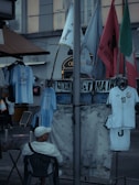 A street market scene with various flags and sports jerseys displayed. A man in a white cap and shirt is sitting on a chair facing away from the camera. The jerseys include a well-known football player's name. Several flags are visible, including green, red, and blue ones, and there is a floral-patterned cloth hanging alongside.