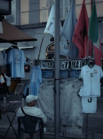 A street market scene with various flags and sports jerseys displayed. A man in a white cap and shirt is sitting on a chair facing away from the camera. The jerseys include a well-known football player's name. Several flags are visible, including green, red, and blue ones, and there is a floral-patterned cloth hanging alongside.