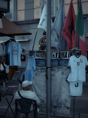 A street market scene with various flags and sports jerseys displayed. A man in a white cap and shirt is sitting on a chair facing away from the camera. The jerseys include a well-known football player's name. Several flags are visible, including green, red, and blue ones, and there is a floral-patterned cloth hanging alongside.