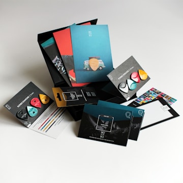 A collection of colorful guitar picks and packaging arranged on a light surface. The packaging features multiple designs and colors, including teal, red, and black, with some picks displayed prominently. One card has an image of a van with a guitar pick on top, and another card shows a close-up of a guitar. Various informational inserts accompany the products, showcasing design and brand details.