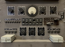 The image features a vintage electronic equipment panel that has multiple knobs, switches, dials, and connectors. Prominent elements include a central analog gauge, labeled with precise measurements, surrounded by adjustment knobs marked for various controls such as timers, rheostats, and other functions. There are several toggle switches and input sockets, along with labeled sections indicating settings and circuitry. The panel is largely industrial gray, giving it a utilitarian appearance.