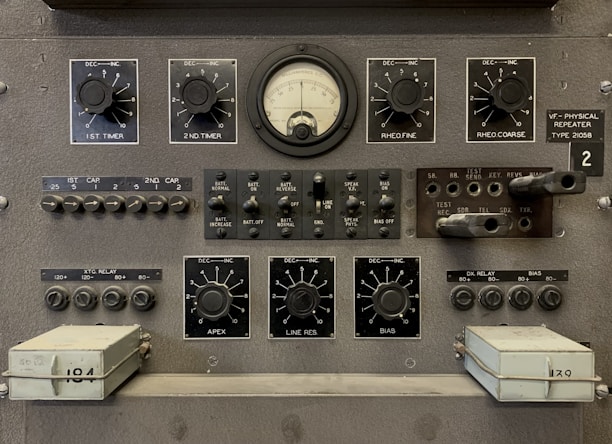 The image features a vintage electronic equipment panel that has multiple knobs, switches, dials, and connectors. Prominent elements include a central analog gauge, labeled with precise measurements, surrounded by adjustment knobs marked for various controls such as timers, rheostats, and other functions. There are several toggle switches and input sockets, along with labeled sections indicating settings and circuitry. The panel is largely industrial gray, giving it a utilitarian appearance.
