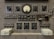 The image features a vintage electronic equipment panel that has multiple knobs, switches, dials, and connectors. Prominent elements include a central analog gauge, labeled with precise measurements, surrounded by adjustment knobs marked for various controls such as timers, rheostats, and other functions. There are several toggle switches and input sockets, along with labeled sections indicating settings and circuitry. The panel is largely industrial gray, giving it a utilitarian appearance.