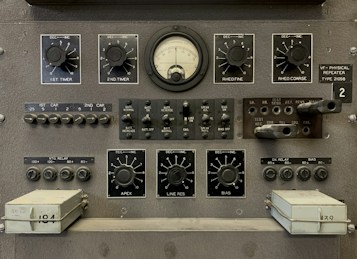 The image features a vintage electronic equipment panel that has multiple knobs, switches, dials, and connectors. Prominent elements include a central analog gauge, labeled with precise measurements, surrounded by adjustment knobs marked for various controls such as timers, rheostats, and other functions. There are several toggle switches and input sockets, along with labeled sections indicating settings and circuitry. The panel is largely industrial gray, giving it a utilitarian appearance.