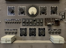 The image features a vintage electronic equipment panel that has multiple knobs, switches, dials, and connectors. Prominent elements include a central analog gauge, labeled with precise measurements, surrounded by adjustment knobs marked for various controls such as timers, rheostats, and other functions. There are several toggle switches and input sockets, along with labeled sections indicating settings and circuitry. The panel is largely industrial gray, giving it a utilitarian appearance.