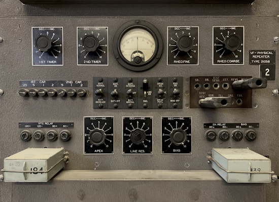 The image features a vintage electronic equipment panel that has multiple knobs, switches, dials, and connectors. Prominent elements include a central analog gauge, labeled with precise measurements, surrounded by adjustment knobs marked for various controls such as timers, rheostats, and other functions. There are several toggle switches and input sockets, along with labeled sections indicating settings and circuitry. The panel is largely industrial gray, giving it a utilitarian appearance.