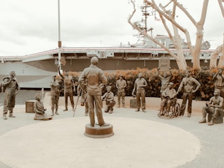 A group of bronze statues depicting military personnel are arranged in a circular formation with a man in an army uniform standing at the center facing them. The figures represent various roles, including pilots, sailors, and other service members, some standing and others seated, one in a wheelchair. In the background, there is a large naval ship, partially obscured by trees with bare branches.