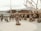 A group of bronze statues depicting military personnel are arranged in a circular formation with a man in an army uniform standing at the center facing them. The figures represent various roles, including pilots, sailors, and other service members, some standing and others seated, one in a wheelchair. In the background, there is a large naval ship, partially obscured by trees with bare branches.