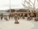 A group of bronze statues depicting military personnel are arranged in a circular formation with a man in an army uniform standing at the center facing them. The figures represent various roles, including pilots, sailors, and other service members, some standing and others seated, one in a wheelchair. In the background, there is a large naval ship, partially obscured by trees with bare branches.