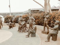 A group of life-sized bronze statues depicting military personnel in various poses. Some are standing, while others are seated or kneeling. The scene includes a figure in a wheelchair, suggesting a theme of camaraderie and inclusion. Behind the statues, there is a naval ship and some trees, adding a maritime and historical context to the scene.