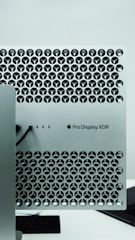 A close-up view of a metallic surface with a lattice pattern of circular cutouts, featuring a Pro Display XDR label and an Apple logo. The design is sleek and modern, typical of high-end electronics.