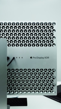 A close-up view of a metallic surface with a lattice pattern of circular cutouts, featuring a Pro Display XDR label and an Apple logo. The design is sleek and modern, typical of high-end electronics.
