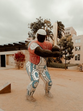 A large, colorful mosaic statue depicting a baseball player holding a bat. The figure is situated outdoors on a sandy path, surrounded by trees and buildings. The statue features intricate patterns and bright colors, with a focus on geometric designs.