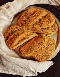 A round loaf of artisanal bread with seeds on top, placed on a gray plate partially covered by a white cloth. The bread has a golden brown color and is sliced, revealing its soft, airy interior.