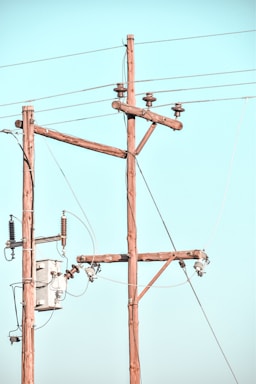 Wooden utility poles with cross-arms hold multiple power lines against a clear blue sky. Electrical insulators are attached to the poles, and there is a transformer box visible.
