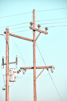 Wooden utility poles with cross-arms hold multiple power lines against a clear blue sky. Electrical insulators are attached to the poles, and there is a transformer box visible.