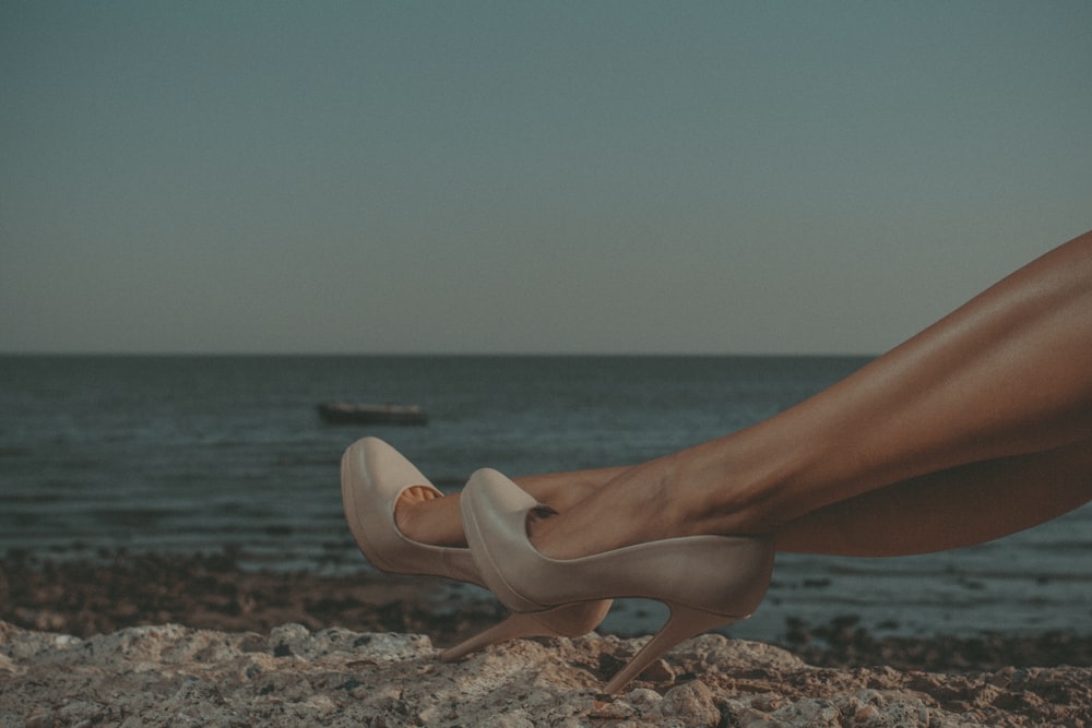 woman in white pumps sitting on brown rock near body of water during daytime