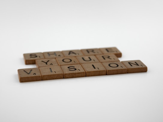 Wooden Scrabble tiles spell out the phrase 'Share Your Vision' against a plain white background. Each tile shows a letter with its corresponding point value, typical of Scrabble games.