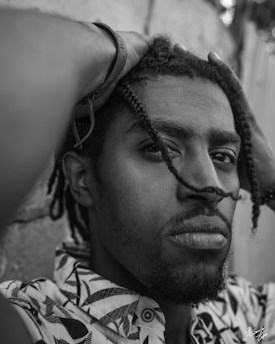 A black and white close-up portrait of a person with braided hair, wearing a patterned shirt. The person is holding their hands on their head, looking directly at the camera with a serious expression. The background is blurred, emphasizing the focus on the subject's face.
