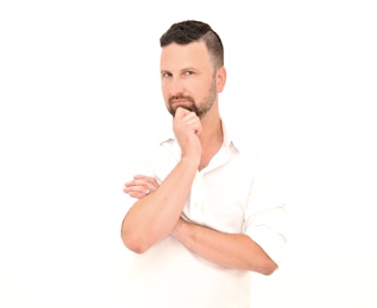 A man with a thoughtful expression, wearing a white shirt, has his hand resting on his chin. The background is completely white, creating a minimalist and clean composition. His hair is short and styled, and he appears to be deeply considering something.