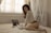 A person is kneeling on a bed, looking at documents in a softly lit room. A laptop is open beside them, and the ambiance is calm and intimate due to the natural light filtering through the blinds.